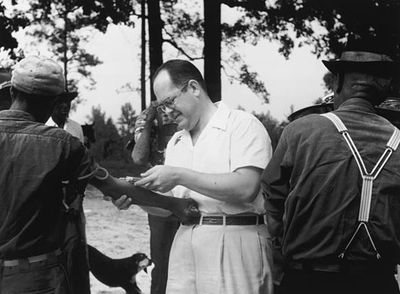 A physician examines a seated Black man outdoors while another man stands nearby.